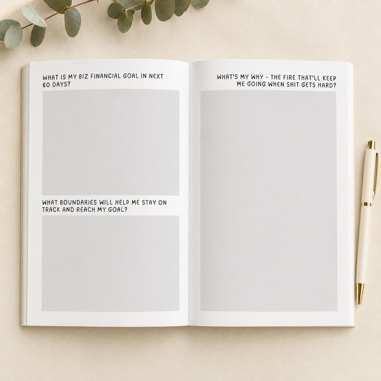 An open notebook or journal spread on a light beige surface, with printed introspective prompts on the left page and blank writing areas on the right. A cream pen with a gold tip rests on the right side, and eucalyptus leaves are visible at the top edge.