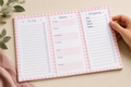 A three-panel pink gingham weekly planner notepad is laid flat on a beige surface, with a hand holding the right panel open. The notepad includes sections for To Do, Menu, and Shopping, with lined spaces and checkboxes.