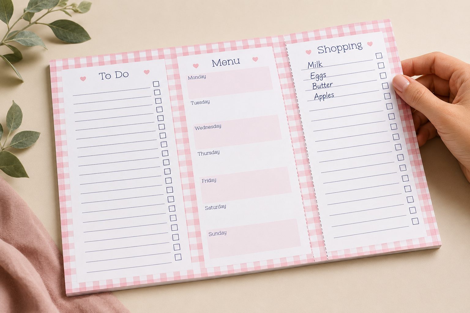 A three-panel pink gingham weekly planner notepad is laid flat on a beige surface, with a hand holding the right panel open. The notepad includes sections for To Do, Menu, and Shopping, with lined spaces and checkboxes.
