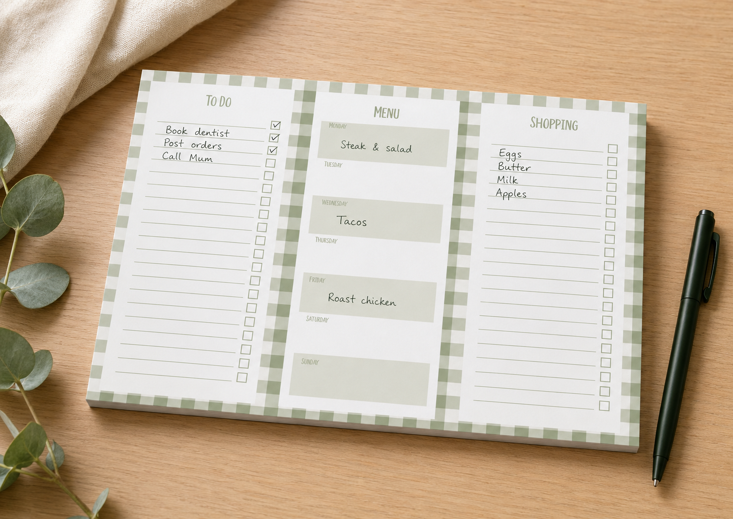 A three-panel weekly planner notepad with a green and white gingham border, displayed on a light wood tabletop alongside a black pen, a beige fabric corner, and eucalyptus leaves. The center panel is labeled 'MENU' with sections for lunch, dinner, and snacks, while the left and right panels show 'To Do' and 'Shopping' lists.