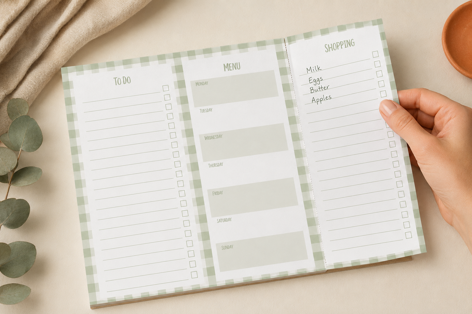 A printable family to-do list and shopping planner card styled in a flat-lay on a beige fabric background with eucalyptus leaves and a wooden slice prop. With hand about to tear off page that has a tare line.