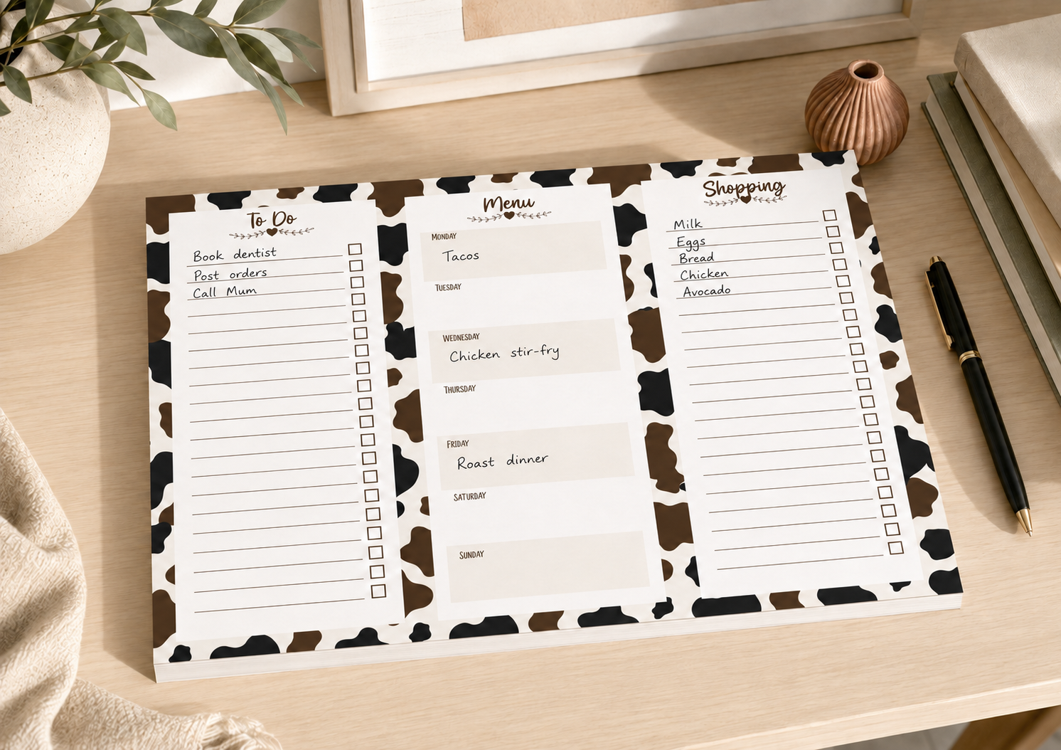 A three-panel weekly planner pad with a black and white cow print border, shown on a light wood desk with a pen, books, and a plant nearby.