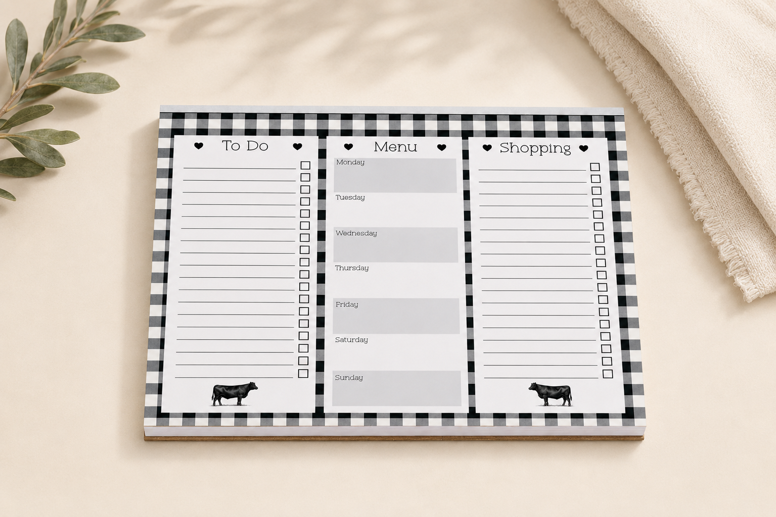 A rectangular notepad with a black-and-white gingham border and three columns labeled To Do, Menu, and Shopping, styled on a beige surface with a folded cream textile and green leaves in the corners.