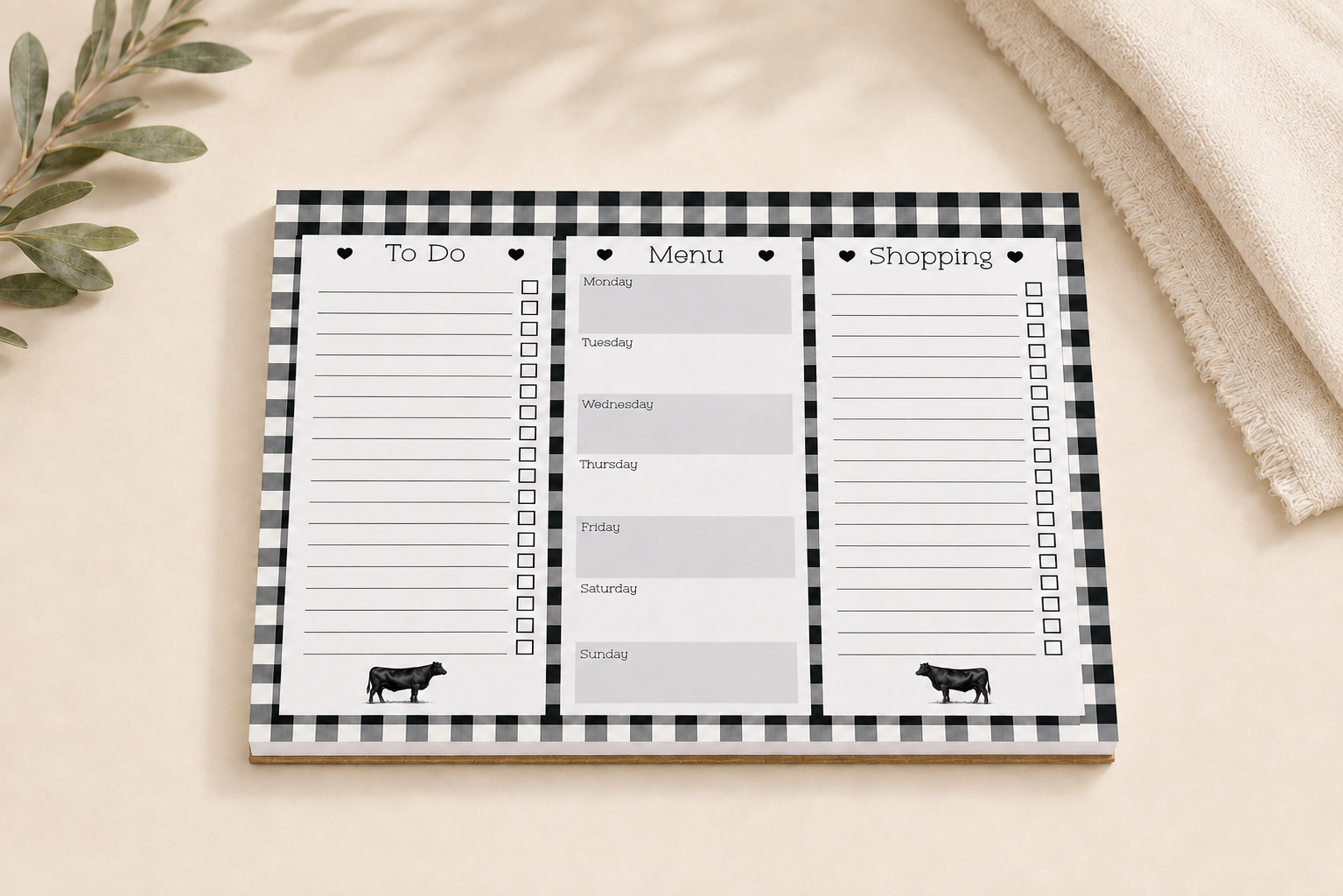 A black and white gingham notepad with three columns labeled To Do, Menu, and Shopping, styled on a beige surface with a soft blanket and eucalyptus leaves.
