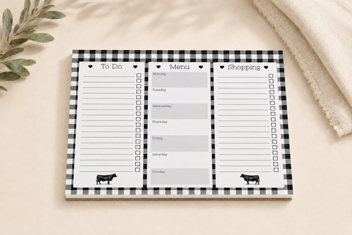 A black and white gingham notepad with three columns labeled To Do, Menu, and Shopping, styled on a beige surface with a soft blanket and eucalyptus leaves.