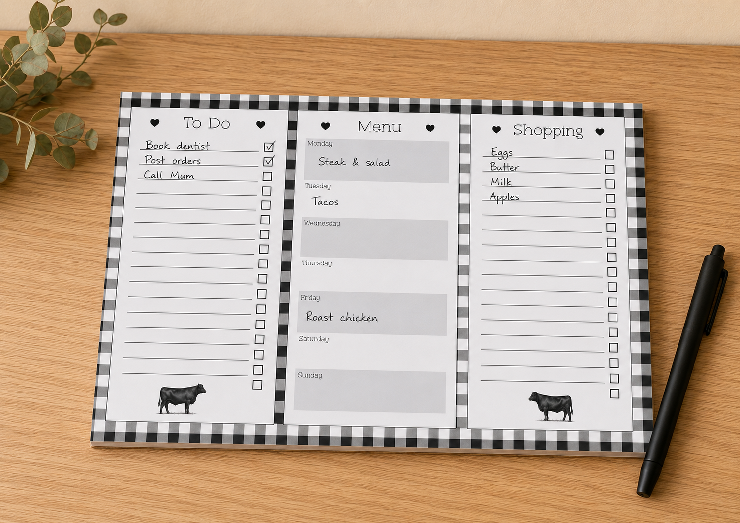 A three-panel weekly planner pad with a black and white gingham border, shown on a light wood tabletop with a pen and small plant sprig nearby.