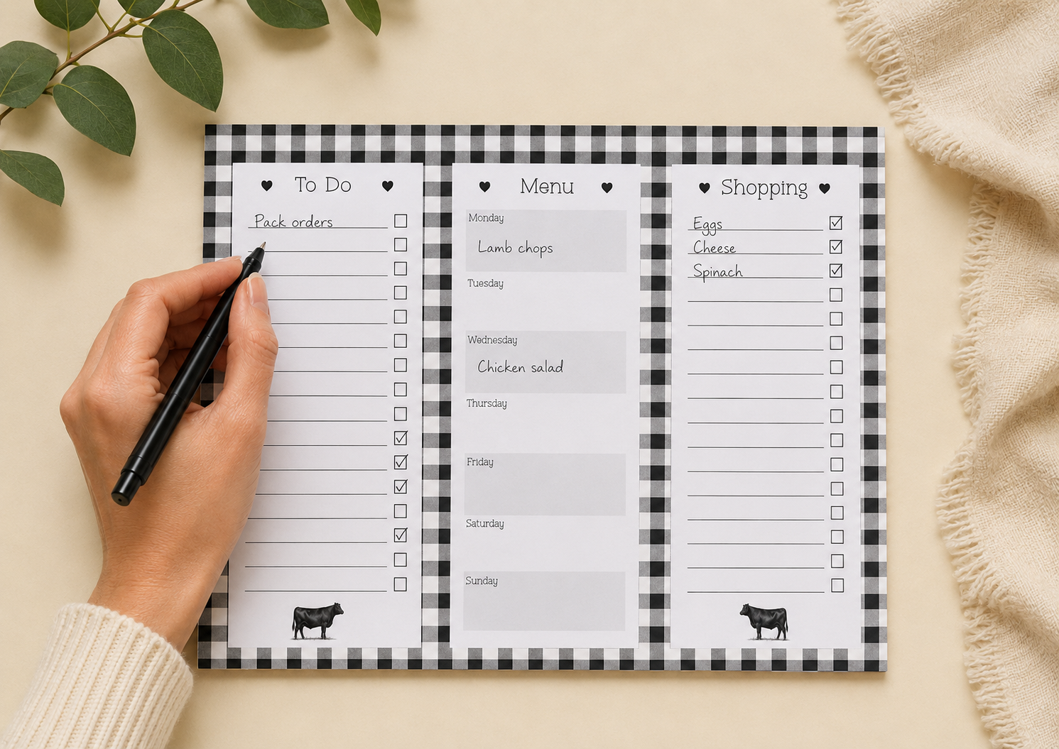 A three-panel checklist planner pad with a black-and-white gingham border, shown on a beige surface with a hand holding a black pen. The planner includes sections labeled To Do, Menu, and Shopping, with small cow illustrations at the bottom of each panel.