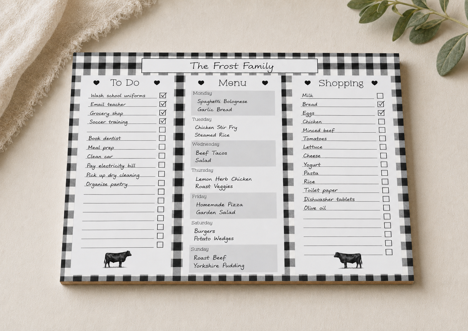 A flat-lay view of a black and white buffalo plaid kitchen planner pad titled 'The Frost Family'. The pad features three columns for 'To Do', 'Menu', and 'Shopping' with checkboxes and placeholder text.