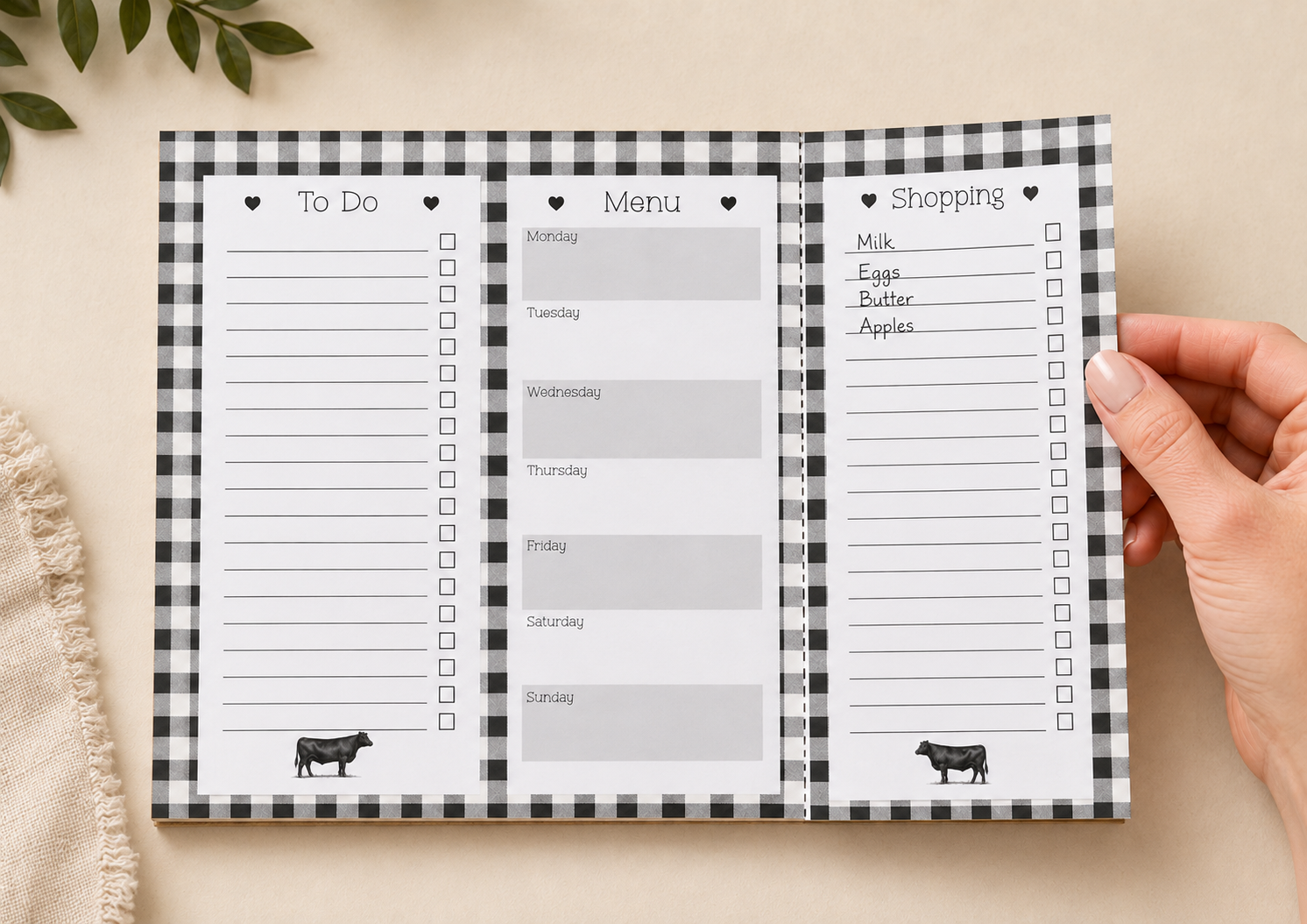A three-panel weekly planner spread with a black-and-white gingham border, shown open on a beige surface. A hand is holding the right panel, and the planner includes sections for To Do, Menu, and Shopping, with small cow illustrations at the bottom of the outer panels.