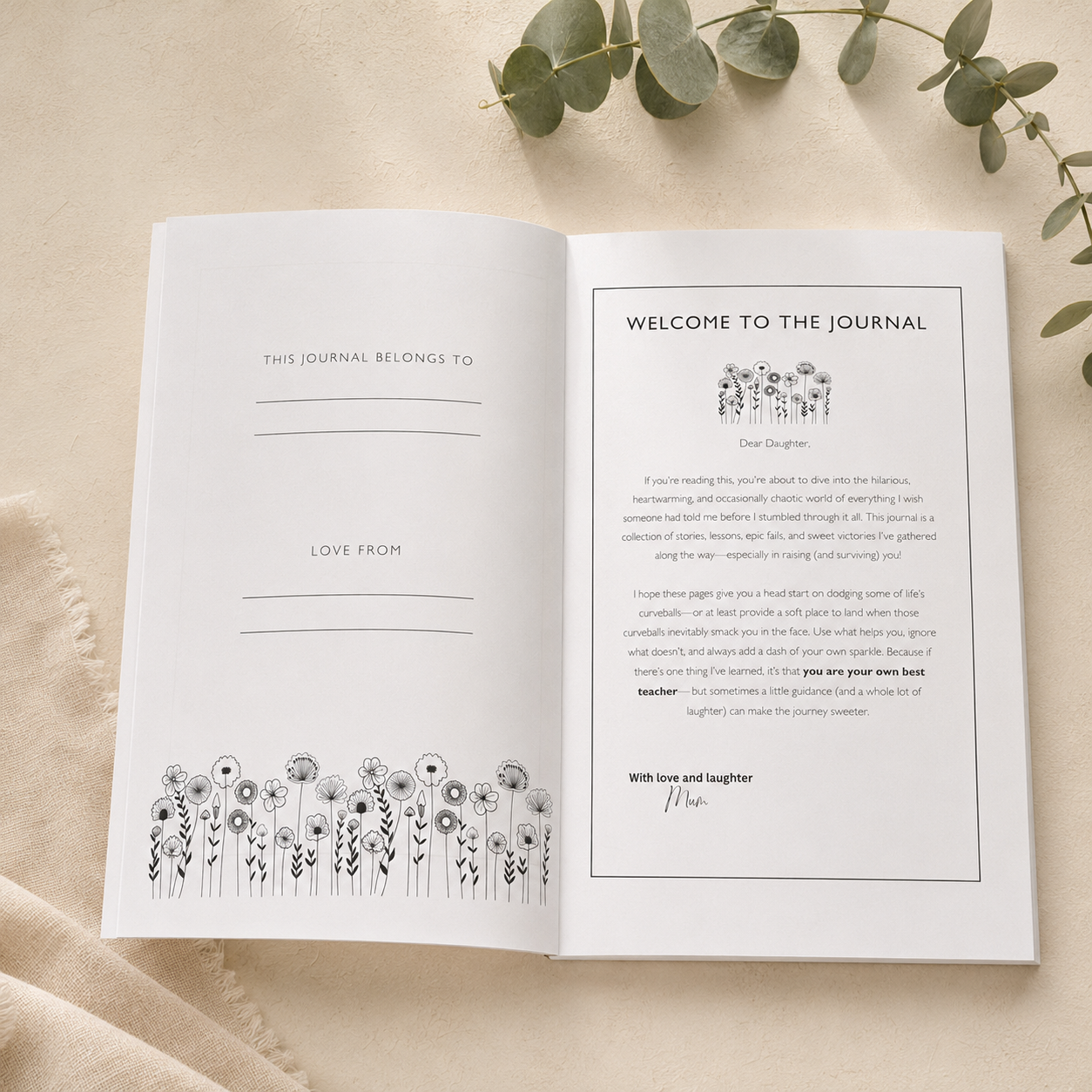 An open journal laid flat on a light beige fabric surface with eucalyptus leaves at the top. The left page is blank with lines and the text 'THIS JOURNAL BELONGS TO' and 'LOVE FROM,' while the right page shows a welcome letter titled 'WELCOME TO THE JOURNAL' with black line illustrations.