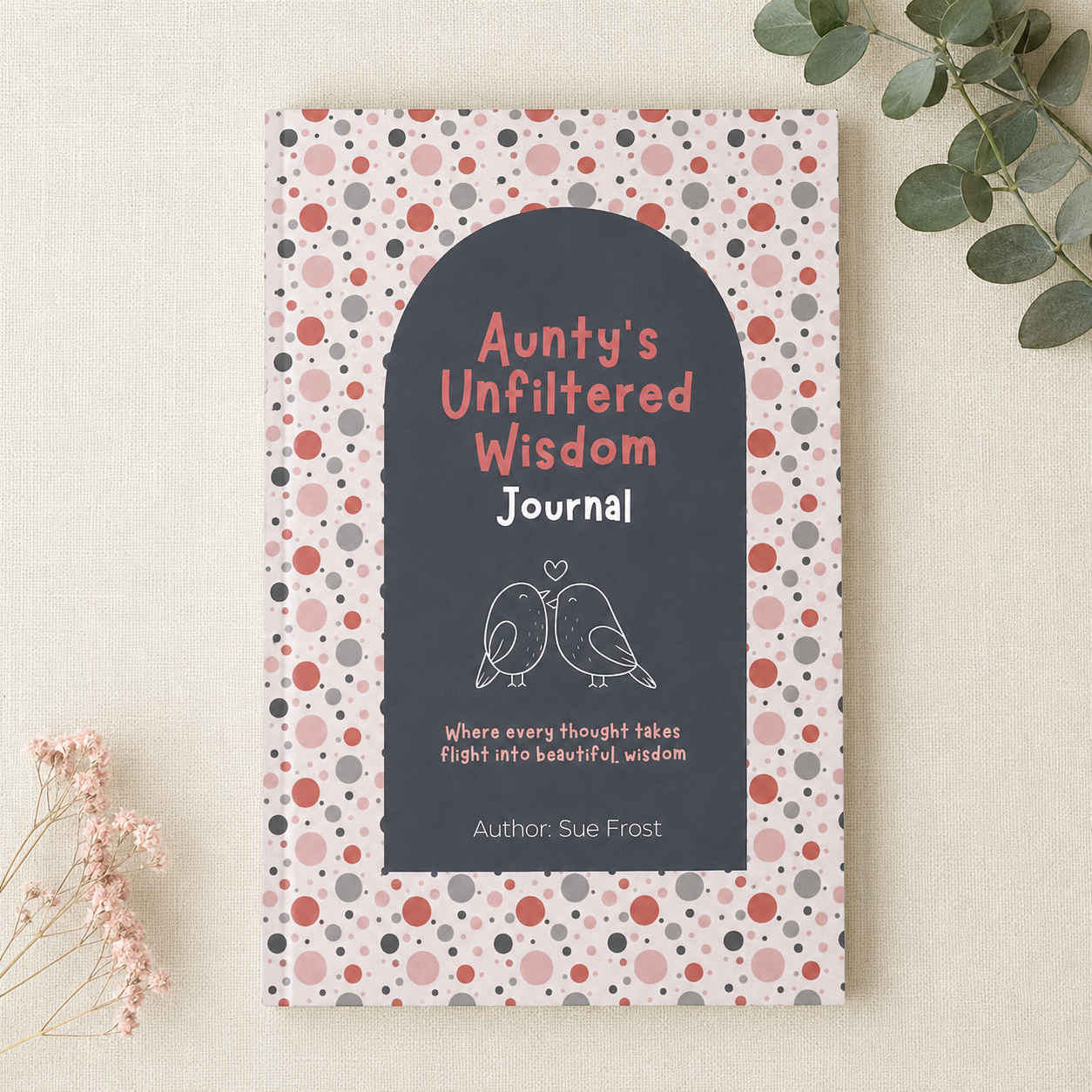 A hardcover journal titled 'Aunty's Unfiltered Wisdom Journal' by Sue Frost, featuring a dark gray arched center with white and pink text and bird illustrations, surrounded by a pink, red, and gray polka dot patterned cover. The book is photographed flat on a light beige textured background with decorative dried flowers and green leaves in the corners.