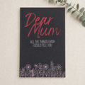 A navy blue hardcover notebook with 'Dear Mum' written in red script and 'ALL THE THINGS I WISH I COULD TELL YOU' in white text, decorated with a floral border at the bottom, displayed on a textured beige linen background with a green eucalyptus sprig in the corner.