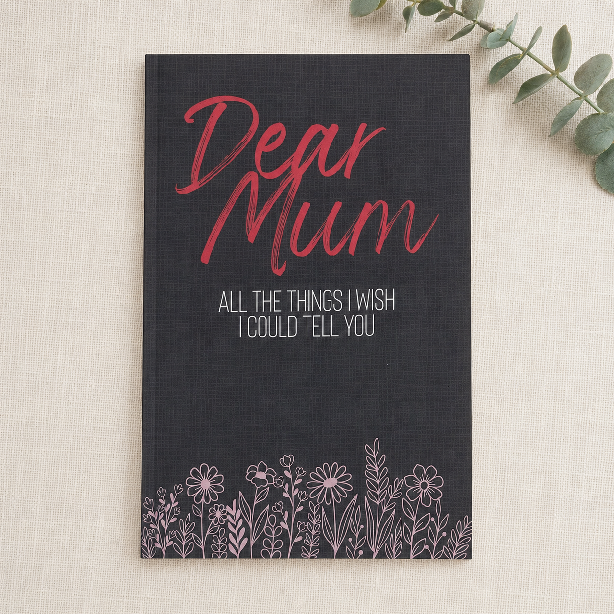 A navy blue hardcover notebook with 'Dear Mum' written in red script and 'ALL THE THINGS I WISH I COULD TELL YOU' in white text, decorated with a floral border at the bottom, displayed on a textured beige linen background with a green eucalyptus sprig in the corner.