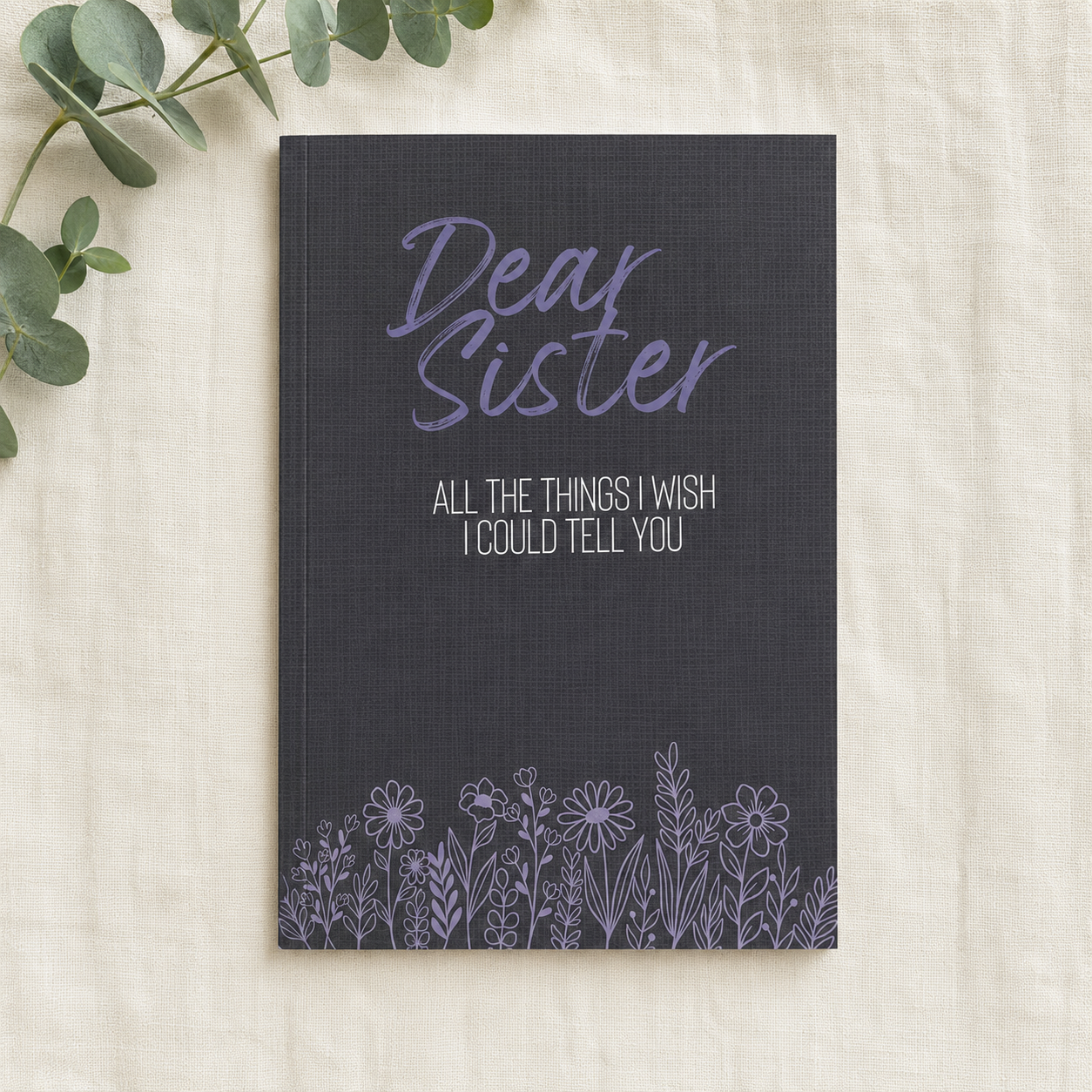 A navy blue hardcover journal with purple and white text reading 'Dear Sister' and 'ALL THE THINGS I WISH I COULD TELL YOU', decorated with a purple floral border at the bottom, displayed on a textured cream fabric with green eucalyptus leaves in the corner.