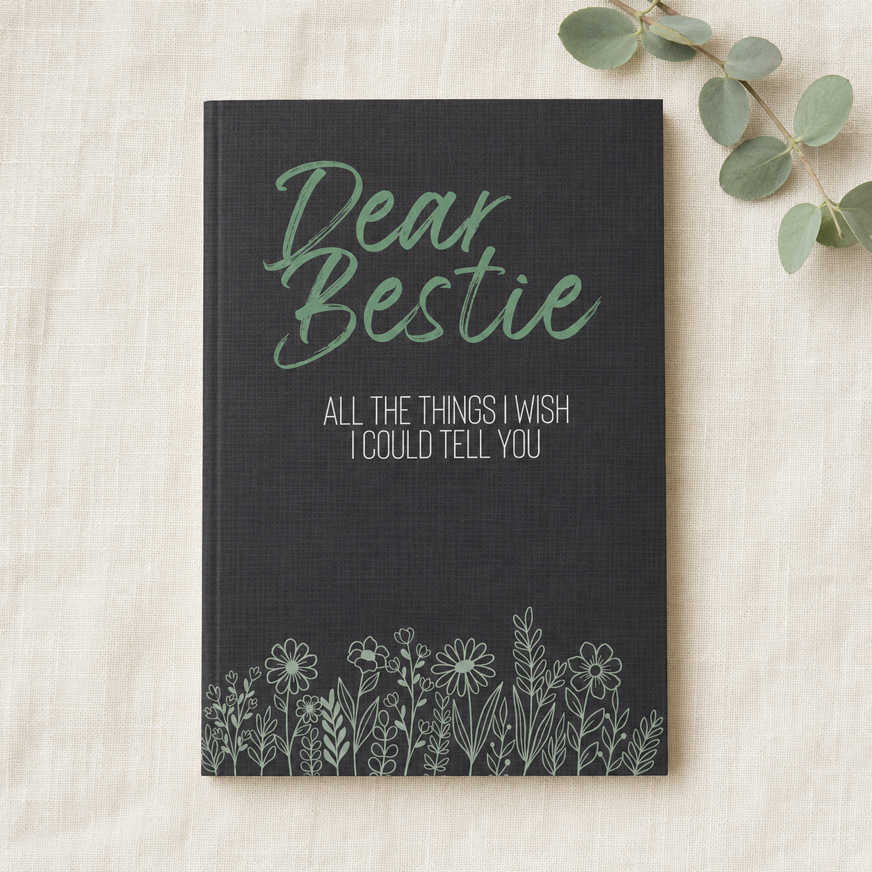 A navy blue hardcover journal with 'Dear Bestie' in sage green script and white text reading 'ALL THE THINGS I WISH I COULD TELL YOU' above a white floral line drawing, styled on a beige fabric background with eucalyptus leaves.