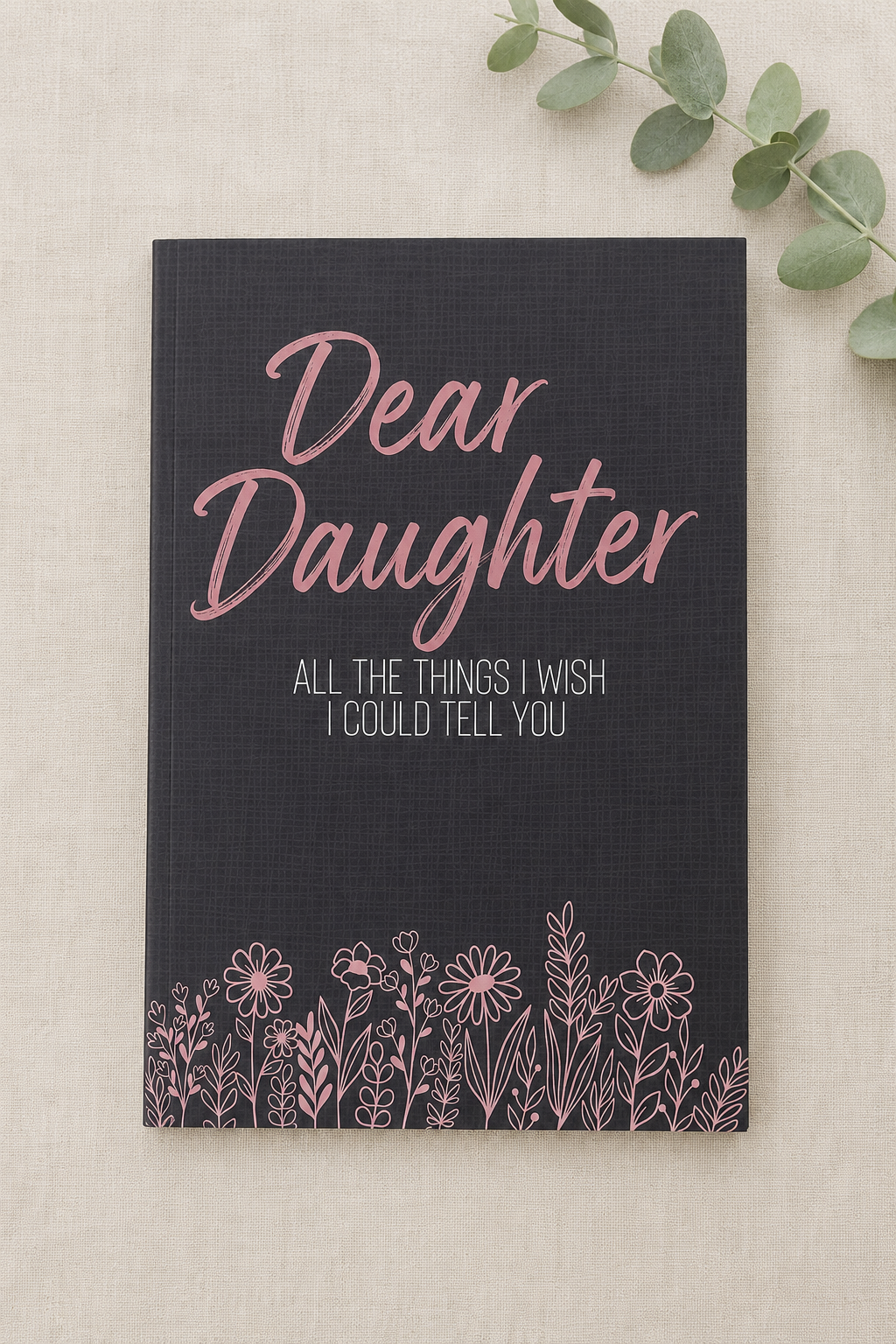 A navy blue hardcover notebook with pink and white text reading 'Dear Daughter' and 'ALL THE THINGS I WISH I COULD TELL YOU,' decorated with a pink floral line-art border at the bottom. It is photographed on a light beige textured fabric background with green eucalyptus leaves in the corner.