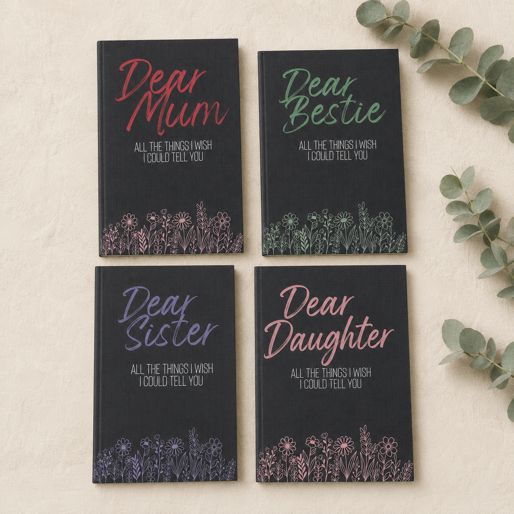 Four hardcover journals arranged in a 2x2 grid on a light beige textured surface with eucalyptus leaves. The covers are navy blue with floral line art at the bottom and titles reading 'Dear Mum', 'Dear Bestie', 'Dear Sister', and 'Dear Daughter' in pink, green, purple, and coral lettering respectively.