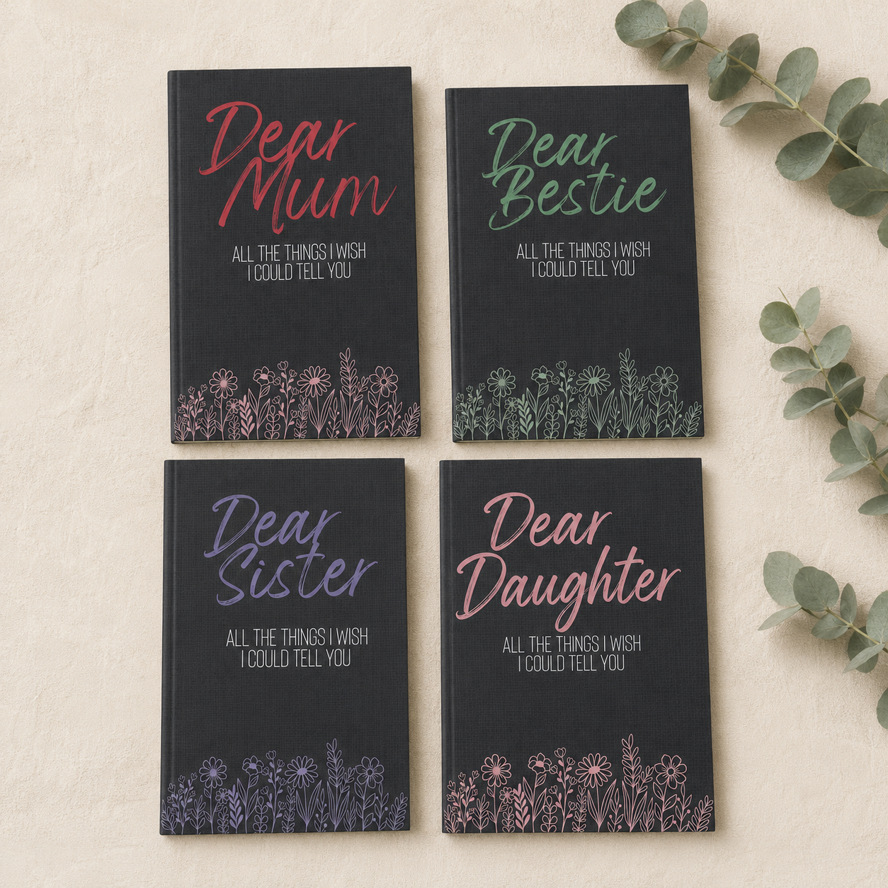 Four hardcover journals arranged in a 2x2 grid on a light beige textured surface with eucalyptus leaves. The covers are navy blue with floral line art at the bottom and titles reading 'Dear Mum', 'Dear Bestie', 'Dear Sister', and 'Dear Daughter' in pink, green, purple, and coral lettering respectively.