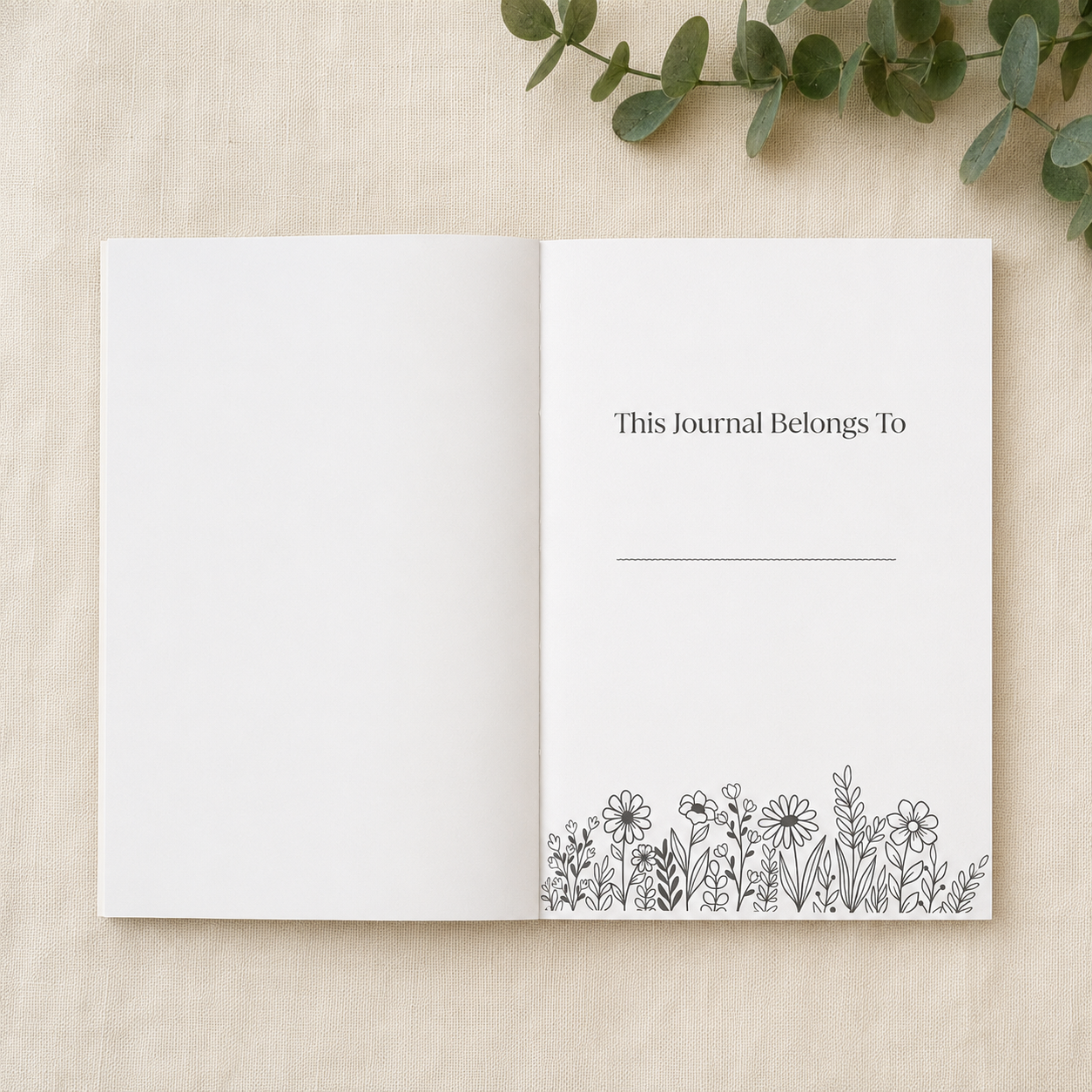 An open blank journal with floral line art at the bottom of the right page, photographed on a light beige fabric background with green eucalyptus leaves in the corner.