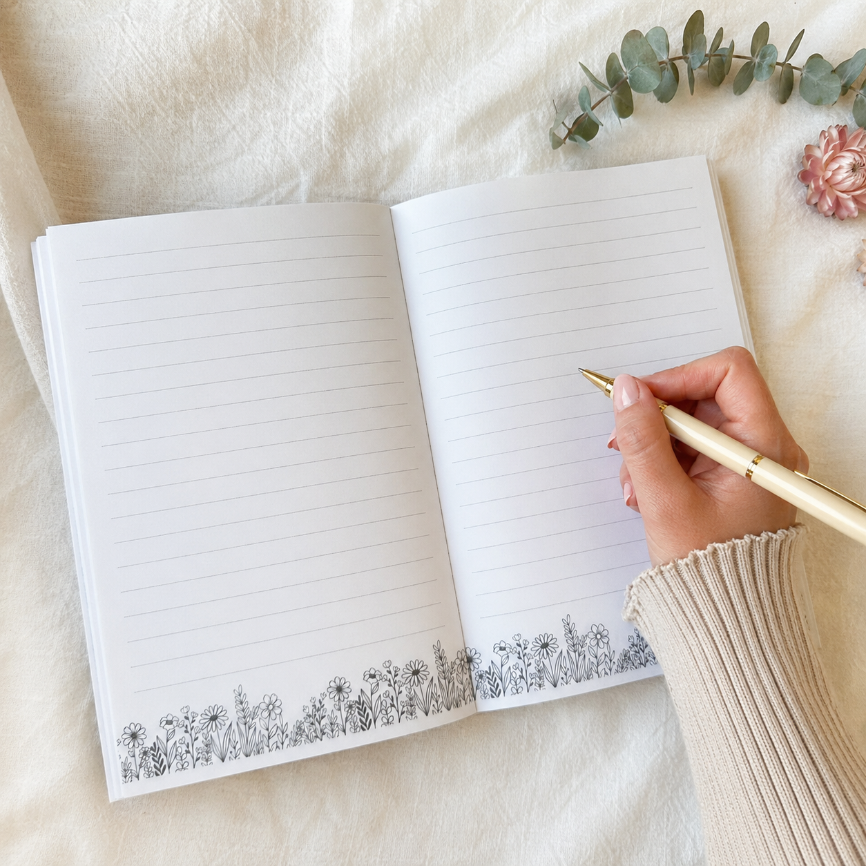 An open lined journal with a black floral border along the bottom, shown on a soft cream fabric background with a hand holding a gold pen.