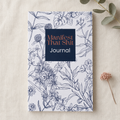 A floral illustrated journal titled 'Manifest That Shit Journal' lies on a light beige fabric surface with small dried botanical props nearby. 