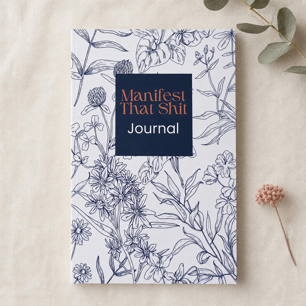 A floral illustrated journal titled 'Manifest That Shit Journal' lies on a light beige fabric surface with small dried botanical props nearby. 