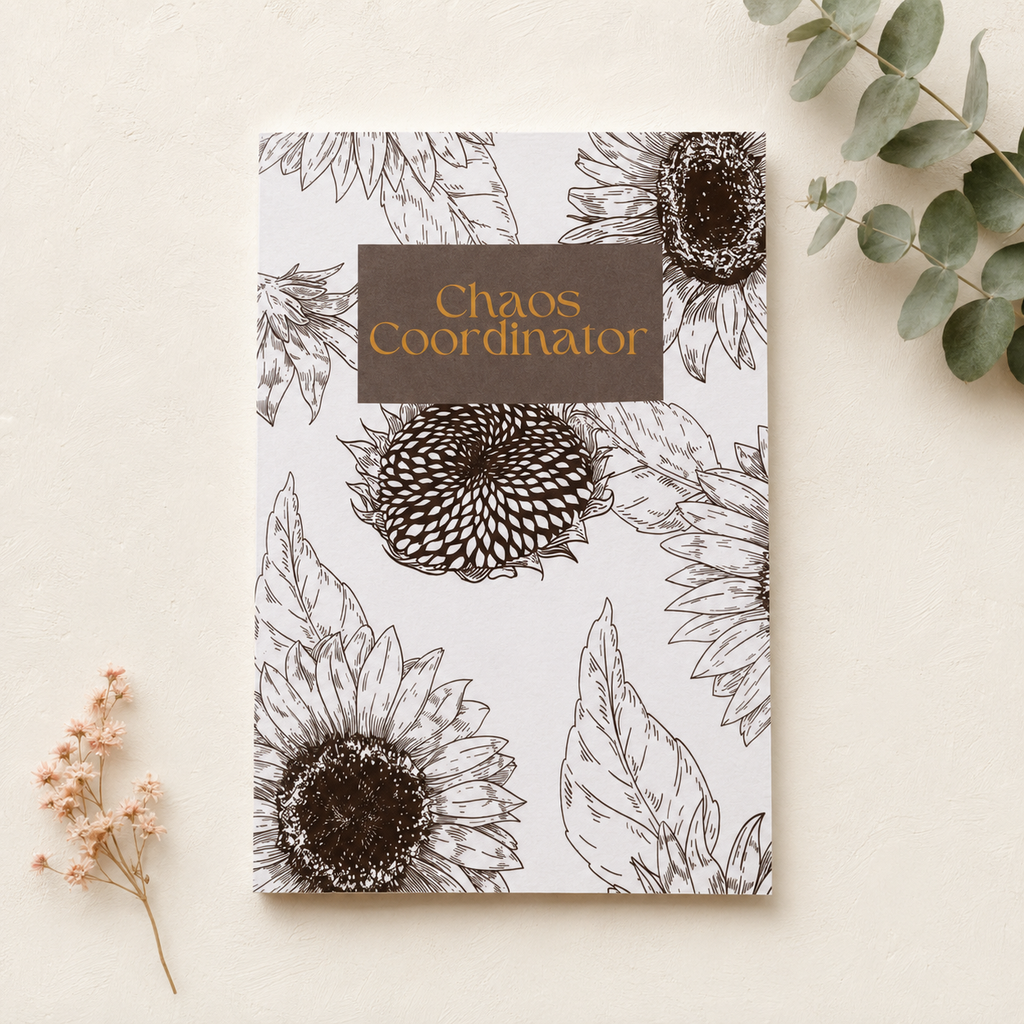 A flat-lay view of a notebook titled 'Chaos Coordinator' with a white cover decorated with black line-art sunflowers and leaves, featuring a brown label with gold text. The notebook is styled on a textured cream background with dried flowers and eucalyptus leaves.