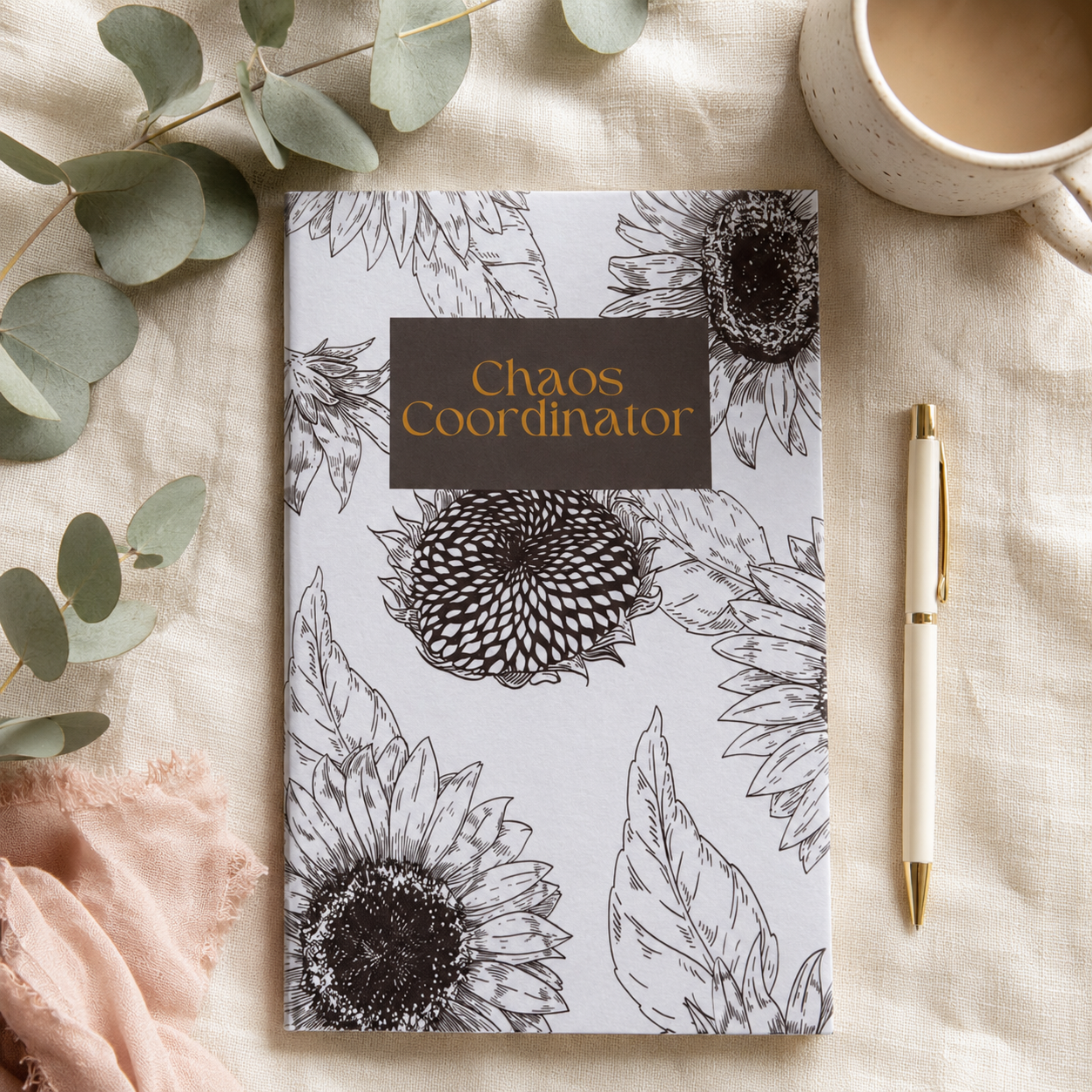 A flat-lay view of a notebook with a white cover decorated with black sunflower illustrations and a dark brown label that reads 'Chaos Coordinator' in gold lettering. The notebook is styled on a beige linen fabric alongside eucalyptus leaves, a pink cloth, a coffee mug, and a gold pen.