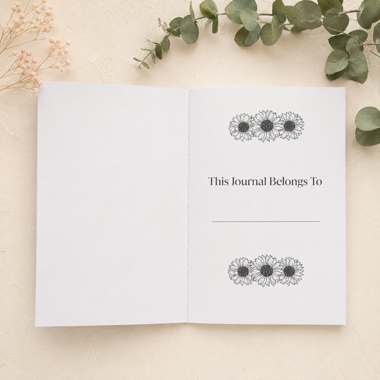 An open white journal or notebook lies on a light beige textured surface, styled with eucalyptus leaves and dried flowers. The right page reads 'This Journal Belongs To' with a blank line below and matching floral motifs near the top and bottom.