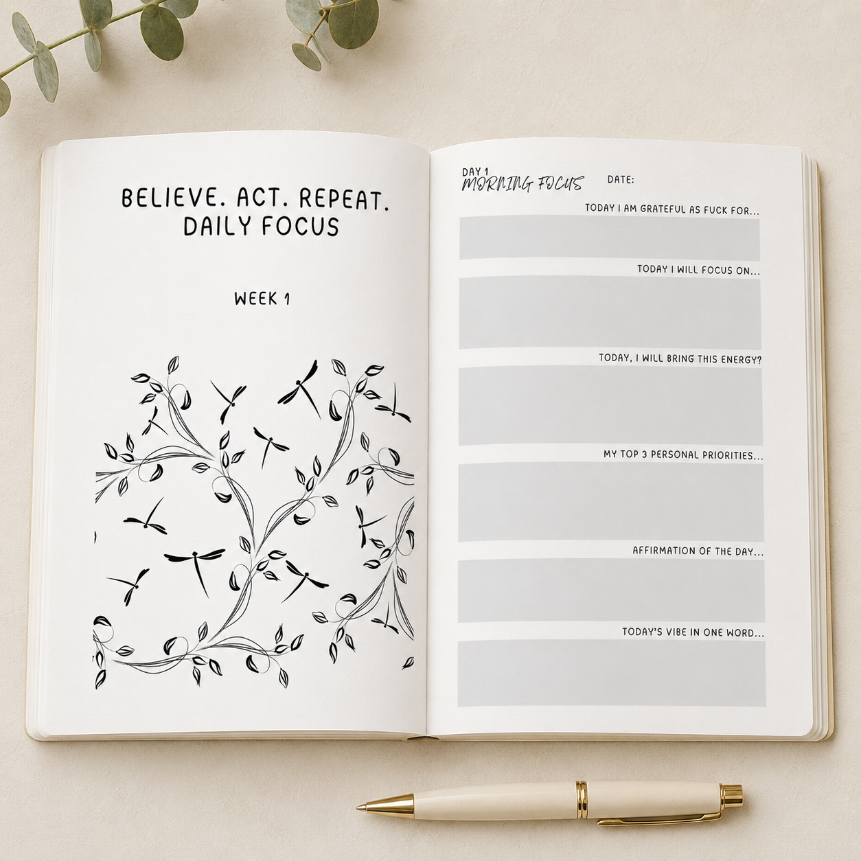 An open daily journal spread showing a 'Daily Focus' page with black text and a black floral vine illustration on the left page, and lined reflection prompts on the right page. A cream-colored pen with gold trim rests below the journal on a light beige surface, with soft green leaves at the top edge.