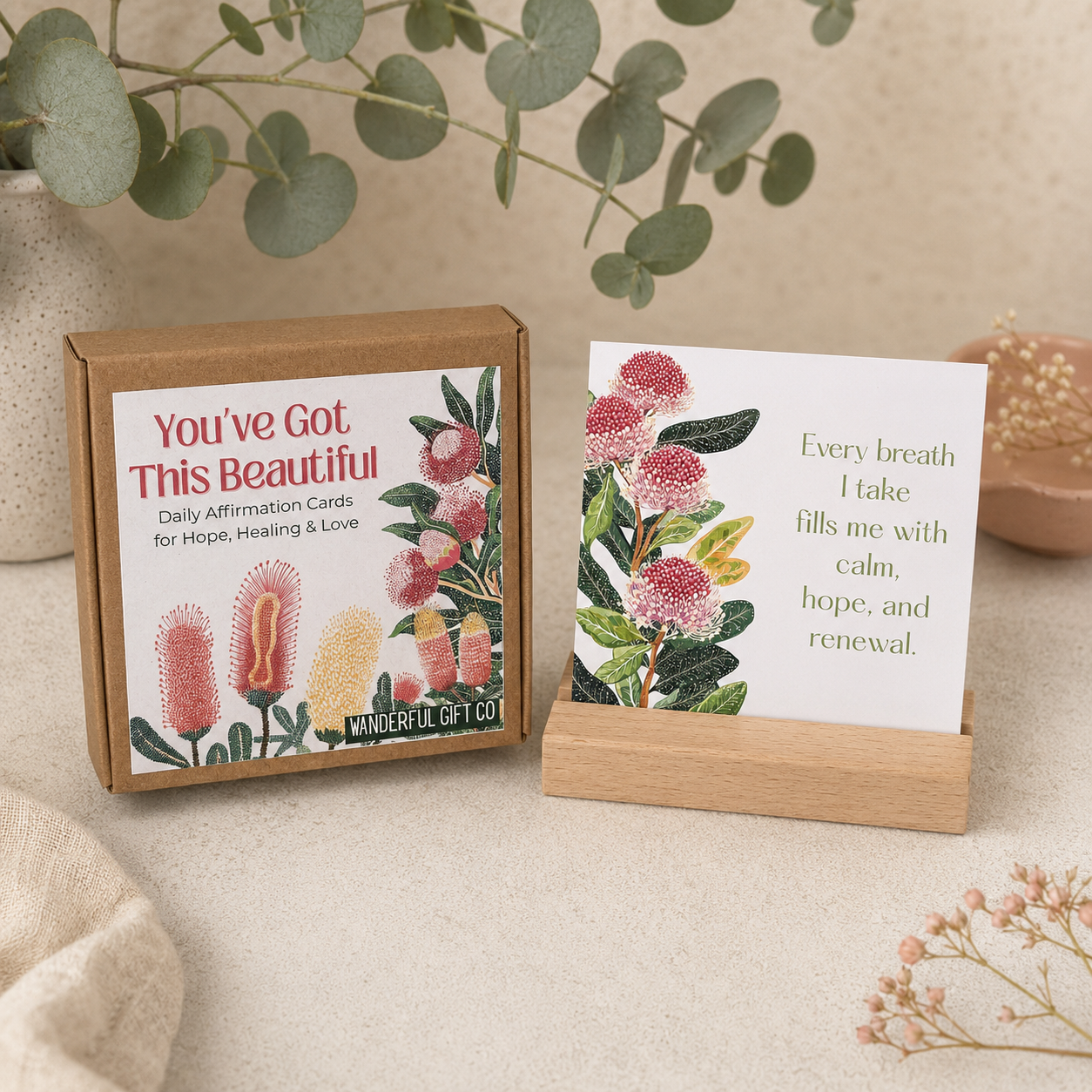 A kraft paper box of affirmation cards sits beside a single card displayed on a small wooden stand. The card has a white background with pink and green botanical illustrations and a quote.