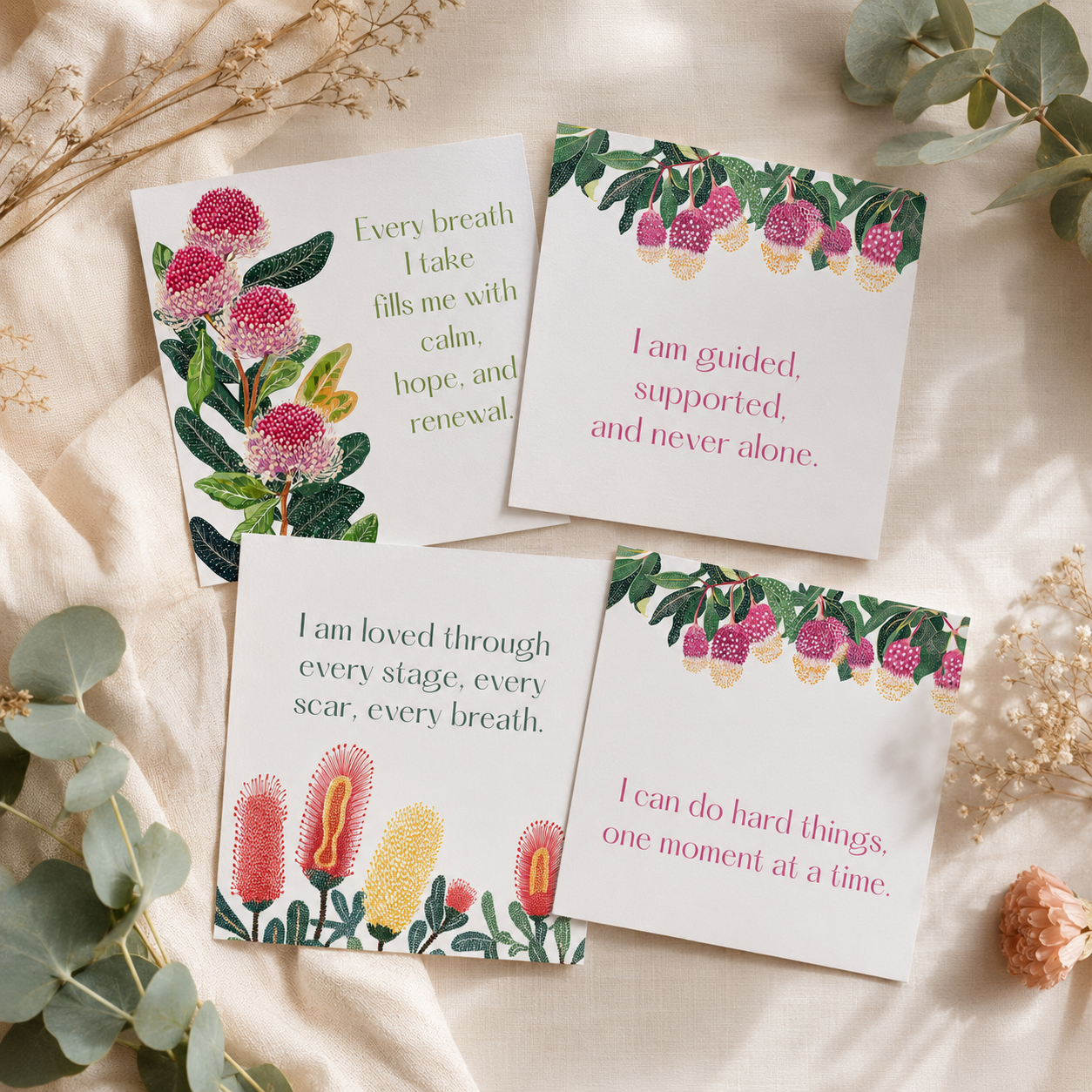Four square printed cards with floral illustrations and inspirational quotes, arranged on a soft cream fabric with dried flowers and greenery as styling props.
