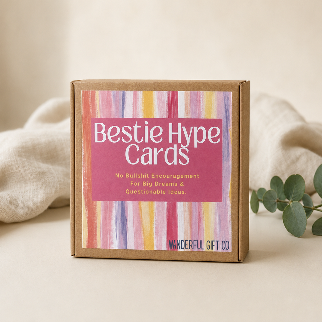A square kraft paper gift box labeled 'Bestie Hype Cards' with a pink label and colorful vertical stripe artwork, styled on a beige surface with soft fabric and greenery in the background.