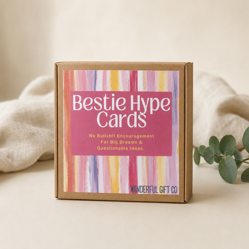 A square kraft paper gift box labeled 'Bestie Hype Cards' with a pink label and colorful vertical stripe artwork, styled on a beige surface with soft fabric and greenery in the background.