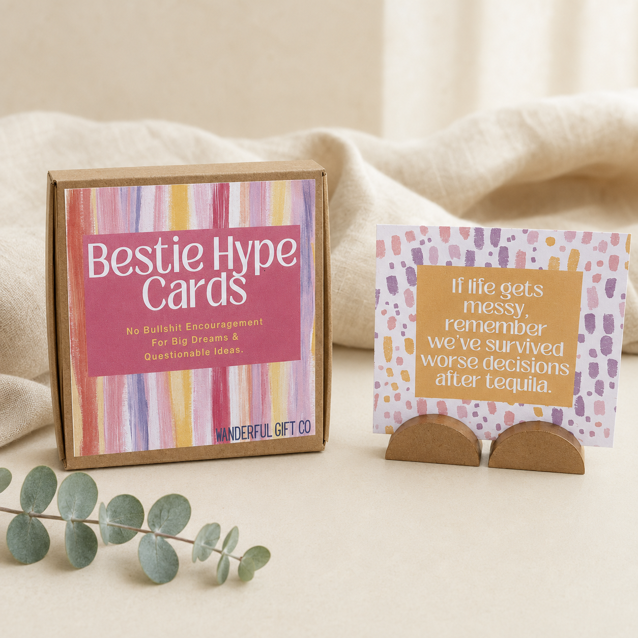 A boxed card product labeled 'Bestie Hype Cards' sits on a soft cream fabric background, with one card displayed on a small stand. The box has a pink front panel with multicolored vertical stripes, and the card has a peach text panel with a white patterned border.