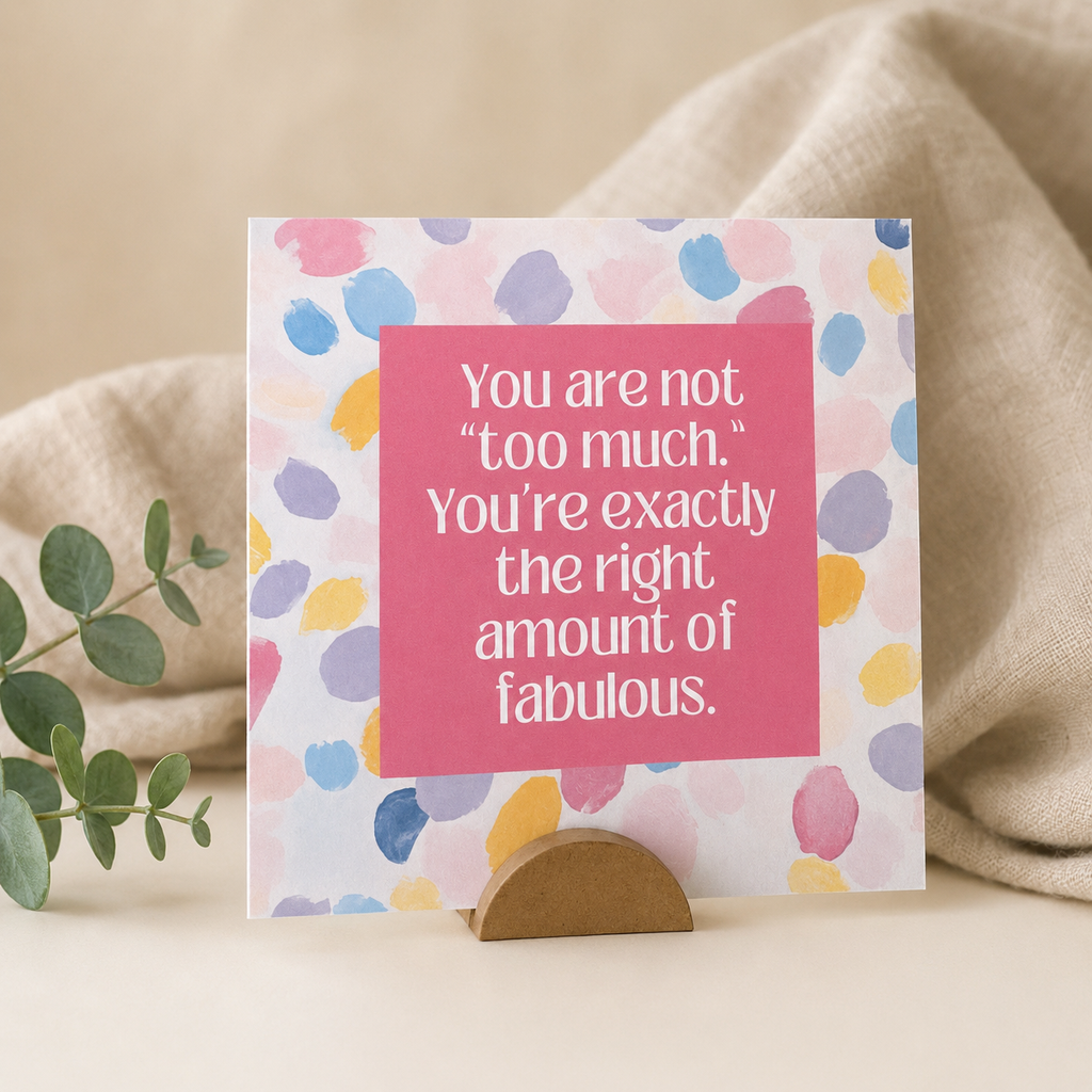 A square greeting card with a pink center panel and multicolored watercolor-style dots on a white background, displayed upright on a small wooden stand with a beige fabric backdrop and a green leaf sprig.