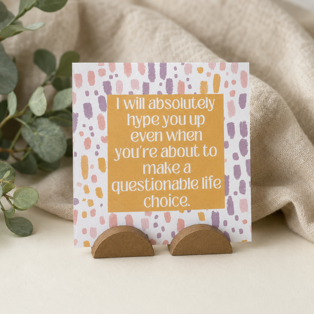 A square greeting card with a patterned border and a central yellow-orange block of text reading, 'I will absolutely hype you up even when you're about to make a questionable life choice.' The card is displayed on a small brown stand against a soft beige fabric backdrop with green leaves at the side.