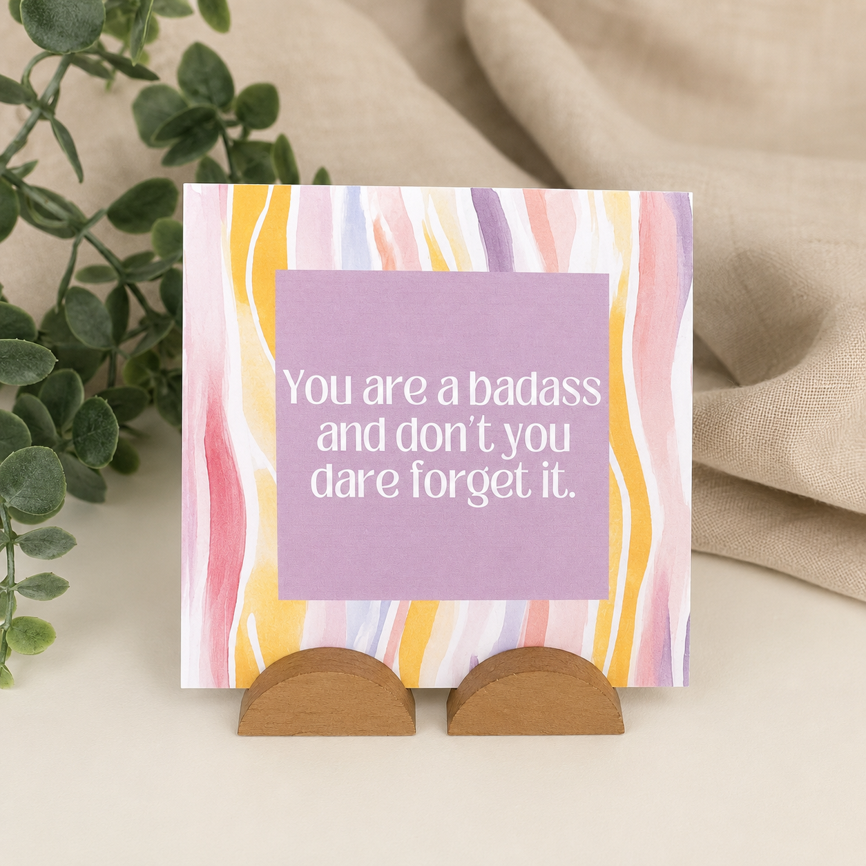 A square greeting card with a pastel multicolor wavy stripe pattern and a lavender center panel reading, 'You are a badass and don't you dare forget it.' The card is displayed on a small wooden stand against a neutral fabric backdrop with green leaves at the side.