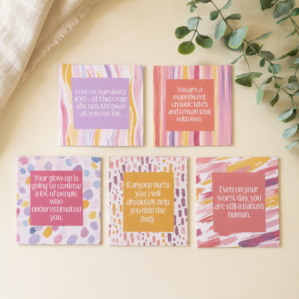 Five colorful greeting cards with humorous and supportive quotes, arranged in a flat-lay on a light beige surface with a textured cream fabric and green eucalyptus leaves.