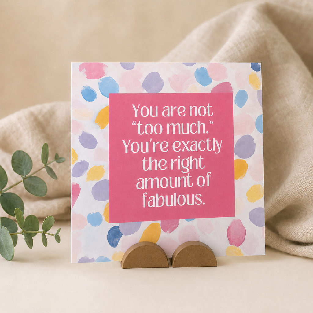 A square greeting card with a pink center panel and multicolored watercolor-style dots on a white background, displayed upright on a small wooden stand with a beige fabric backdrop and a green sprig.