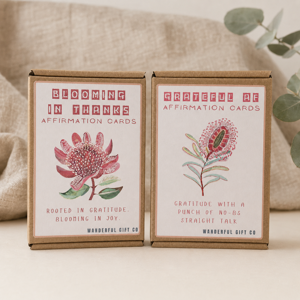 Two brown kraft paper boxes of affirmation cards from Wanderful Gift Co. are displayed side-by-side on a light beige surface with a neutral fabric backdrop and eucalyptus leaves.