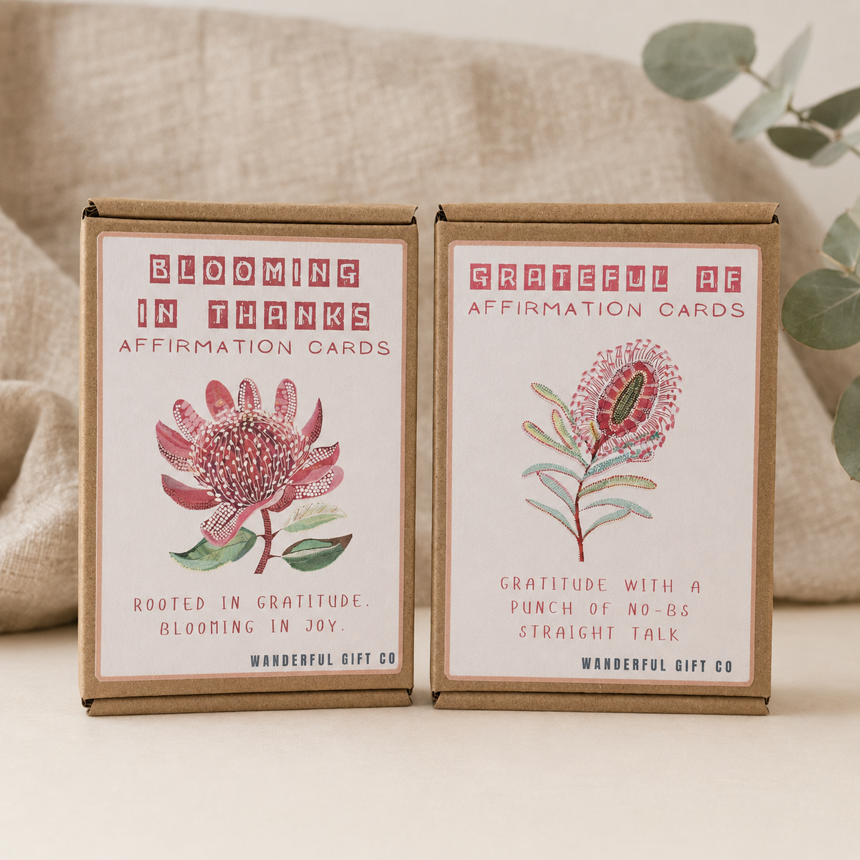 Two brown kraft paper boxes of affirmation cards from Wanderful Gift Co. are displayed side-by-side on a light beige surface with a neutral fabric backdrop and eucalyptus leaves.