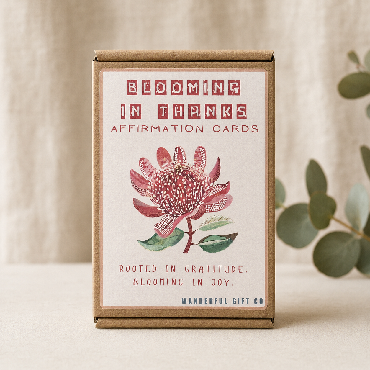 A boxed set of affirmation cards standing upright on a light surface. The package is kraft brown cardboard with a cream front panel featuring the text 'BLOOMING IN THANKS AFFIRMATION CARDS' and an illustration of a pink and green flower.