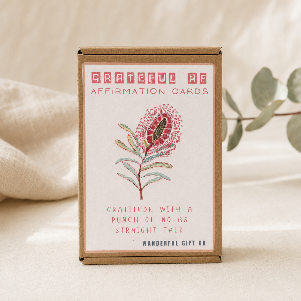 A box of Grateful AF Affirmation Cards by Wanderful Gift Co, featuring a kraft paper box with a white front label that has a pink and green floral illustration and text, set against a soft beige fabric background with blurred eucalyptus leaves.