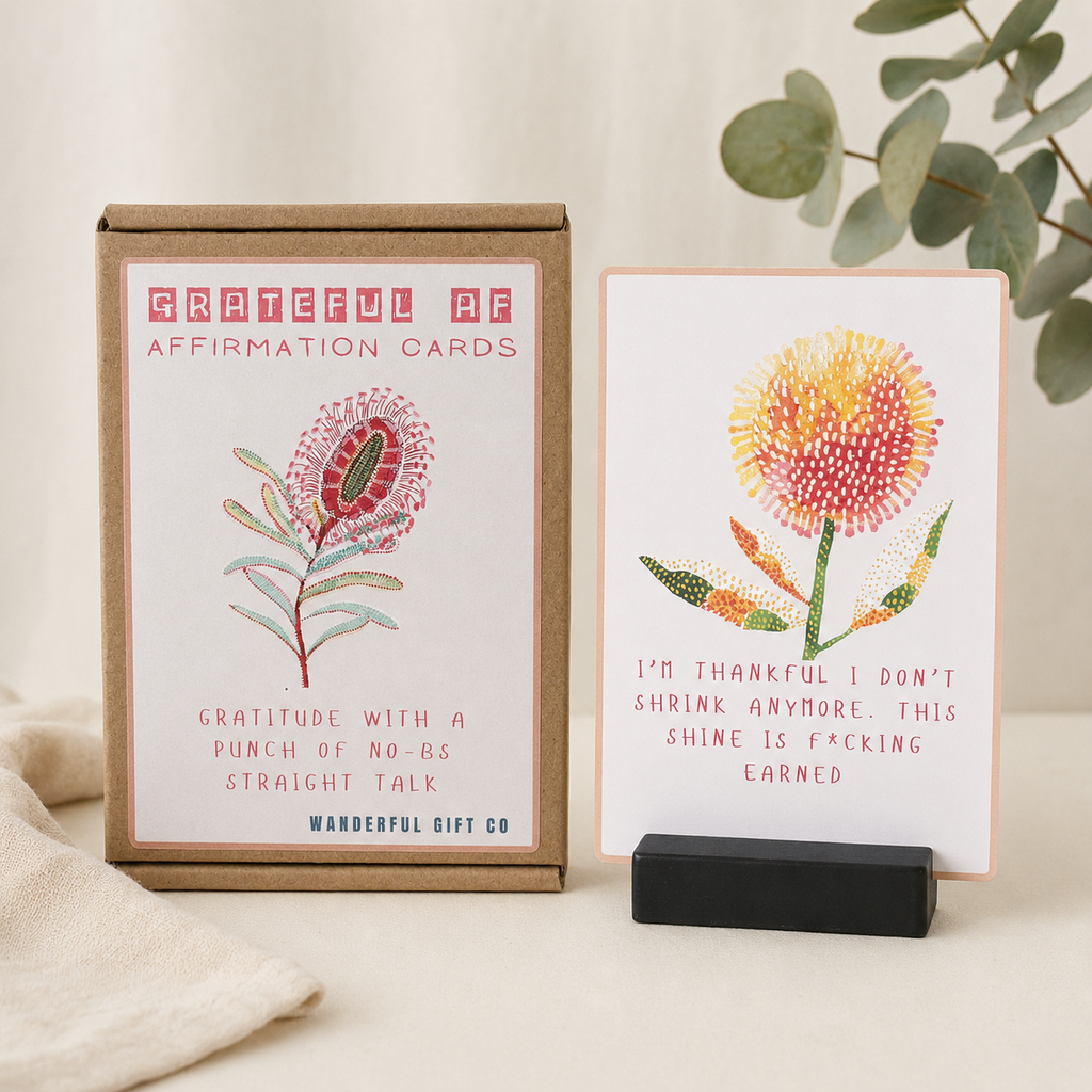 Box of grateful affirmation cards and one sample card displayed on a small black stand, styled on a light surface with fabric and eucalyptus leaves.