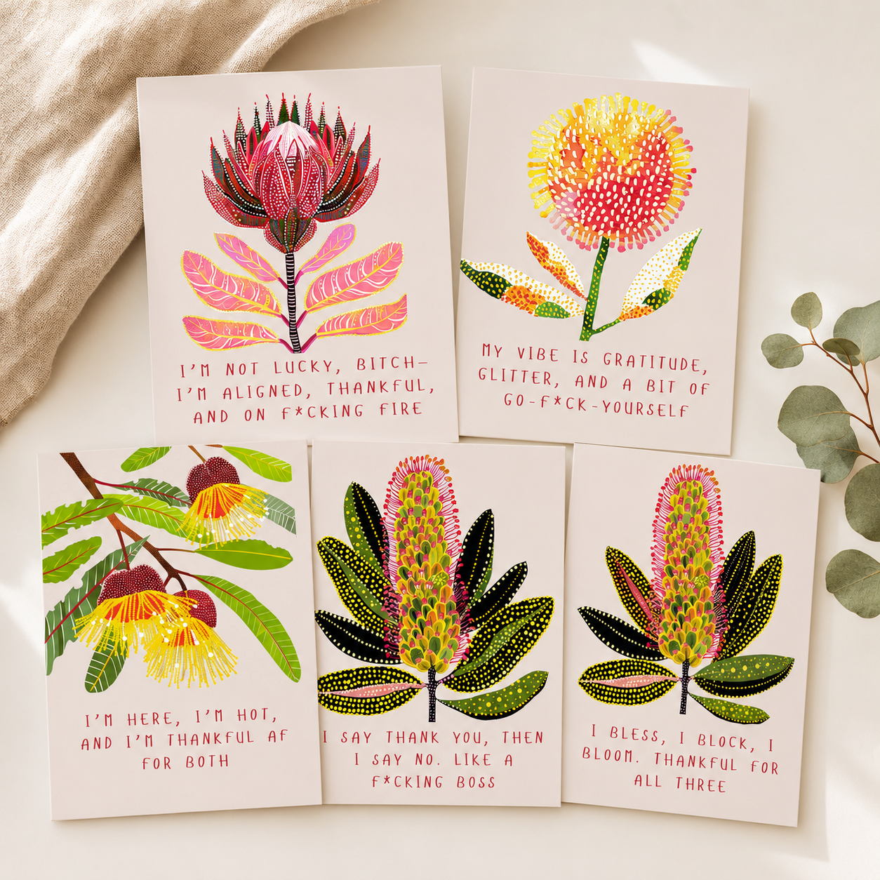 Four botanical greeting cards arranged on a light beige surface with a soft fabric corner and eucalyptus leaves. The cards feature colorful illustrated flowers and humorous text in pink and red lettering.
