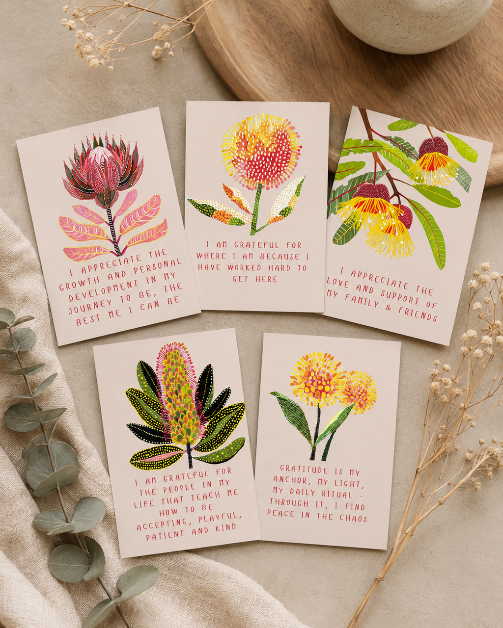 Five botanical greeting cards with floral illustrations and short handwritten-style messages, arranged on a light fabric surface with dried flowers and a ceramic bowl nearby.