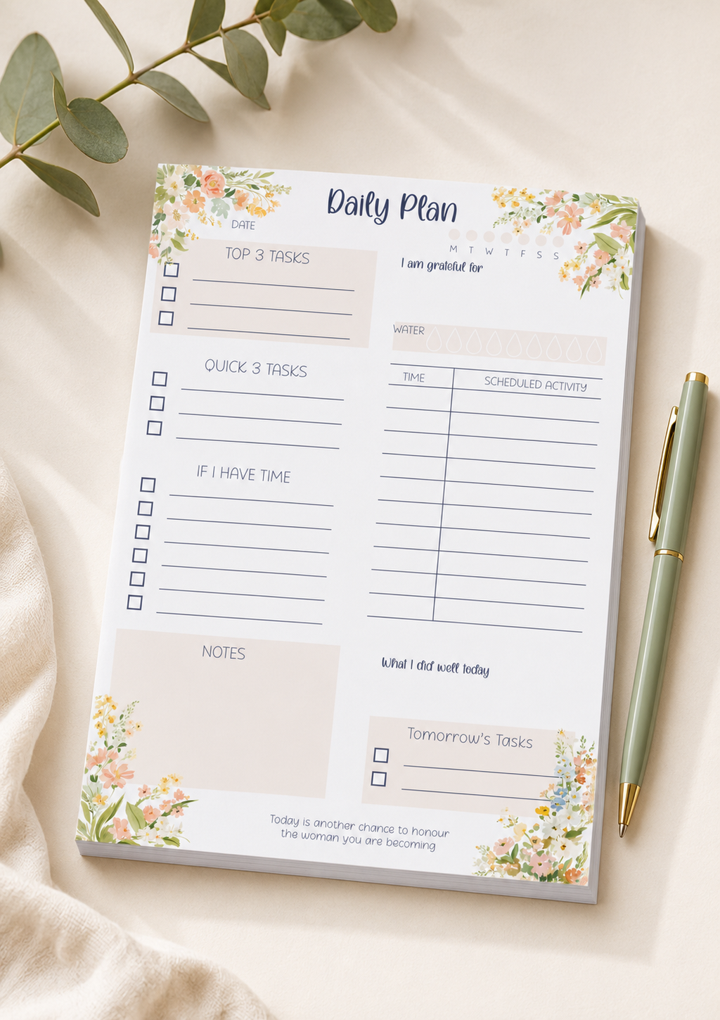 A floral 'Daily Plan' notepad sheet laid flat on a light beige fabric surface, with a green pen beside it and eucalyptus leaves in the corner. The notepad has watercolor-style peach, pink, and green flowers with a cream background and printed task boxes and writing areas.