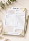 A floral 'Daily Plan' notepad sheet laid flat on a light beige fabric surface, with a green pen beside it and eucalyptus leaves in the corner. The notepad has watercolor-style peach, pink, and green flowers with a cream background and printed task boxes and writing areas.