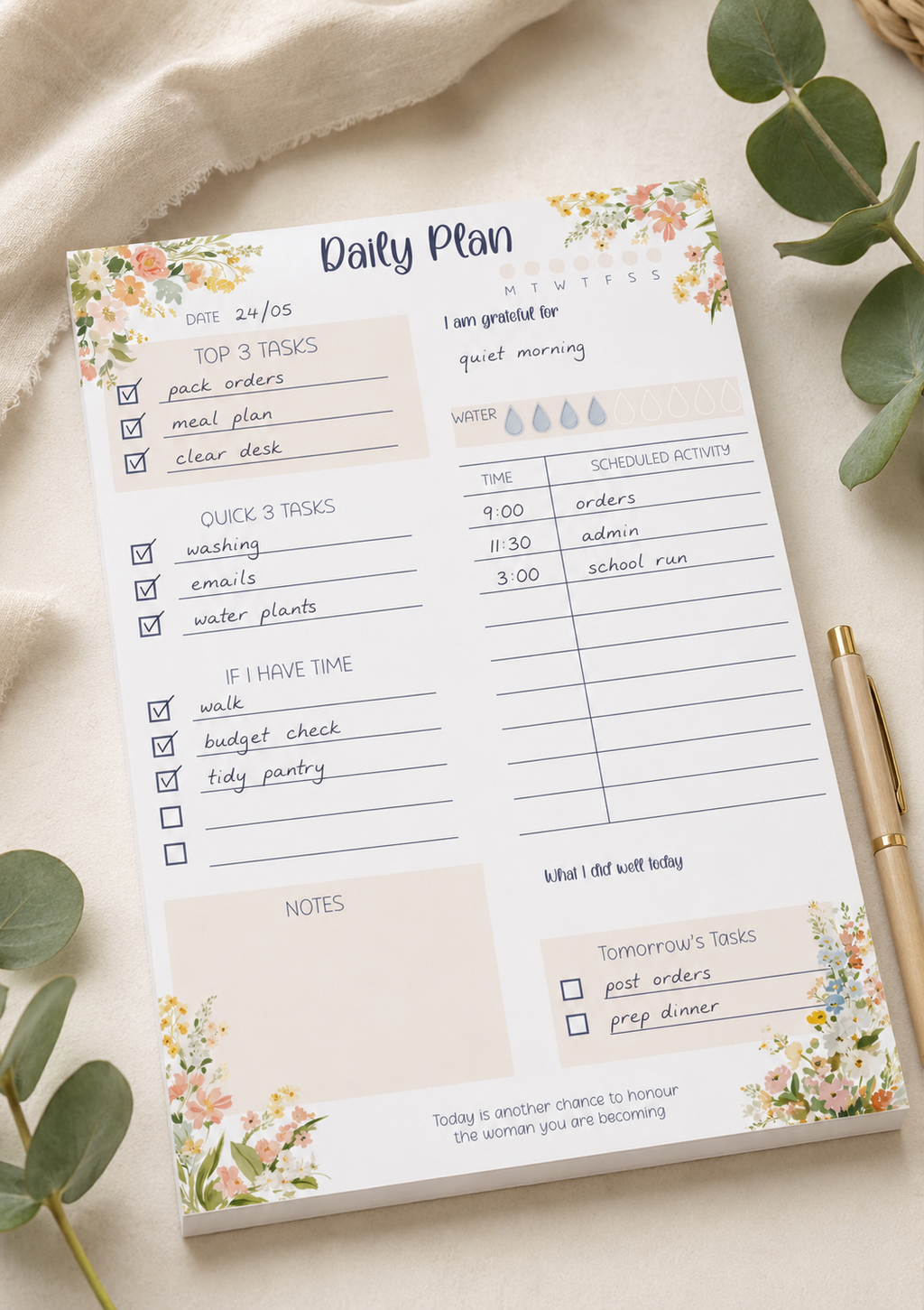 A floral 'Daily Plan' notepad sheet laid flat on a light fabric surface, surrounded by green eucalyptus leaves and a gold pen. The planner page includes sections for tasks, water intake, notes, and tomorrow's tables with pastel watercolor flowers at the corners.
